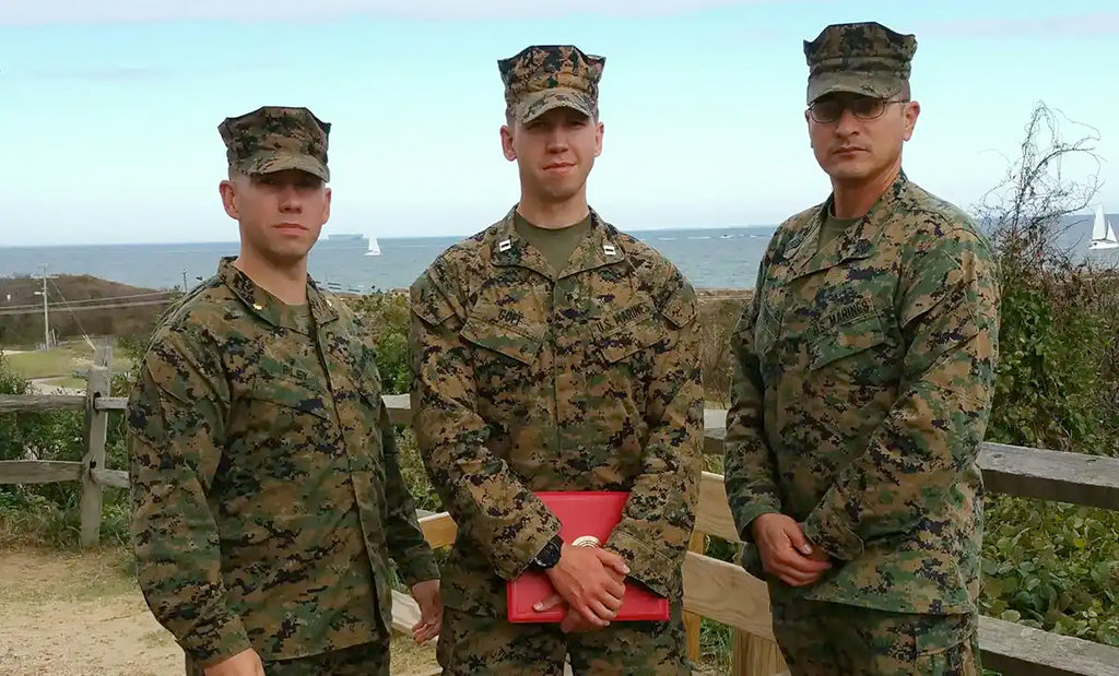 Three Marines stand together in camouflage uniforms, with the ocean in the background. One Marine, in the center, is holding a red folder. The image highlights Jeff Goff, who identified the connection between combat injuries and athletic injuries caused by poorly designed footwear, inspiring his mission to create cleats that support strong, natural arches for optimal performance.